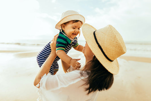 A mama lifting her baby up at the beach, both of them wearing sun hats.